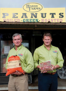 Ken and Brad Hardy pose side-by-side in front of Hardy Farm's peanut stand, toting bushels of boiling peanuts. 